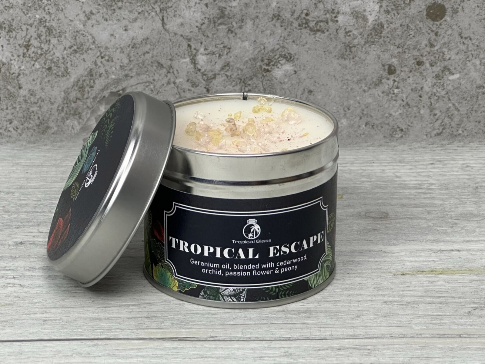 A silver tin candle labeled Tropical Escape sits on a textured gray surface, featuring a design with vibrant tropical plants. The candle is adorned with colorful crystals on top, suggesting a soothing and aromatic experience.