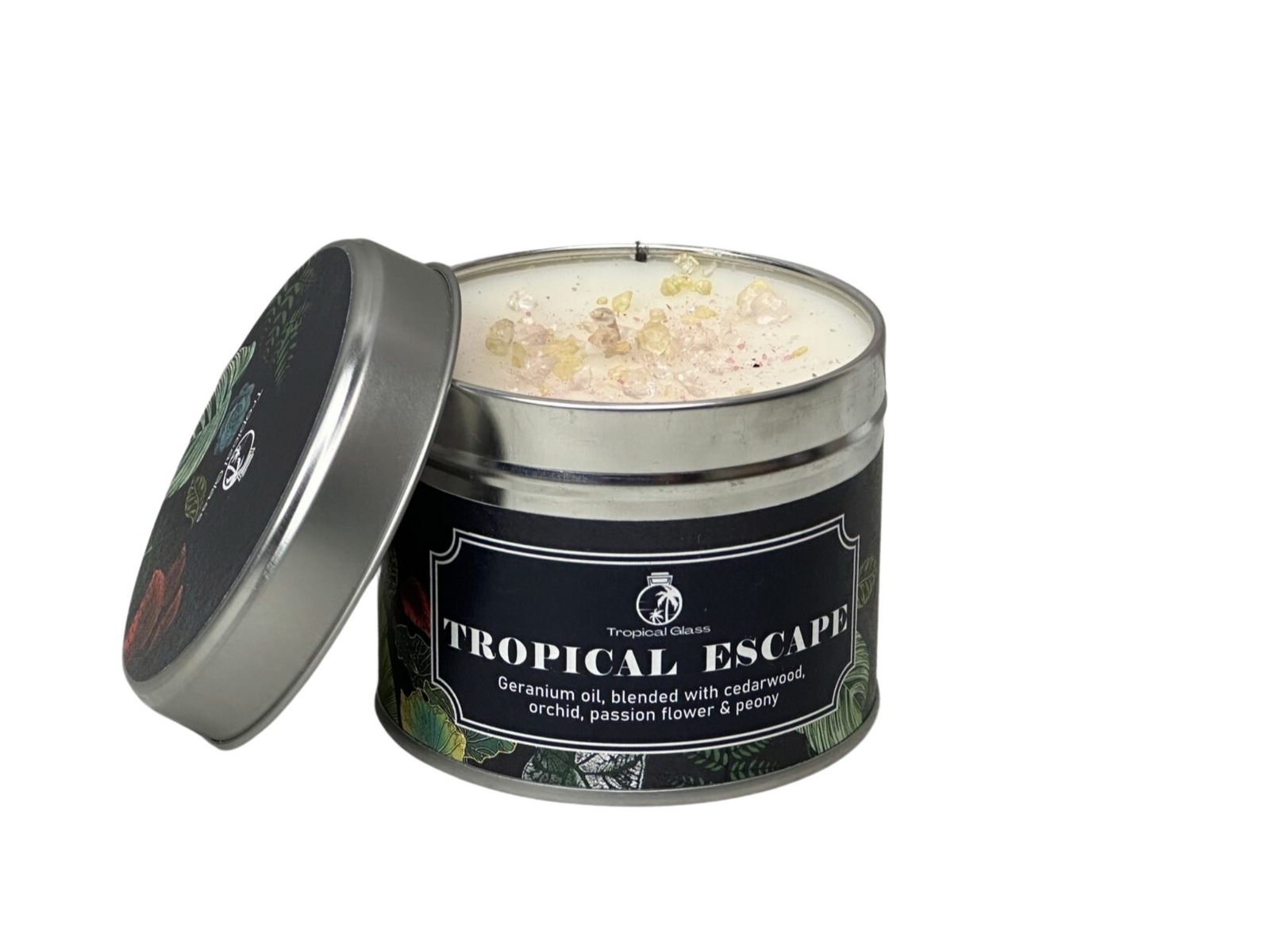 A silver tin candle labeled Tropical Escape features a creamy wax interior adorned with colorful crystals on top. The design incorporates tropical-themed graphics, suggesting a vibrant and exotic ambiance.