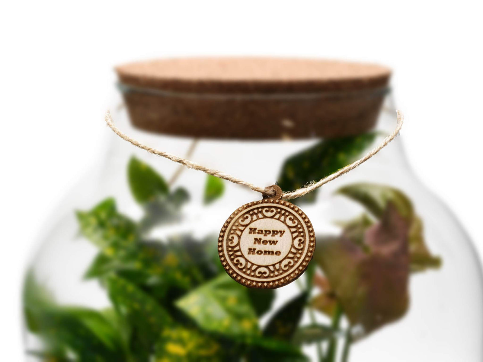 A close-up of a wooden gift tag that reads Happy New Home, attached with twine to a glass jar. The jar contains green plants, adding a natural touch to the display, perfect for celebrating a housewarming occasion.