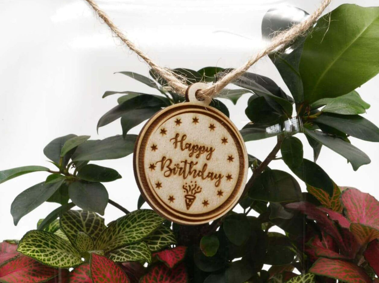 A wooden gift tag with the inscription Happy Birthday is elegantly displayed, surrounded by vibrant green and reddish leaves inside a glass terrarium. The tag is attached with twine, adding a charming touch to the arrangement.