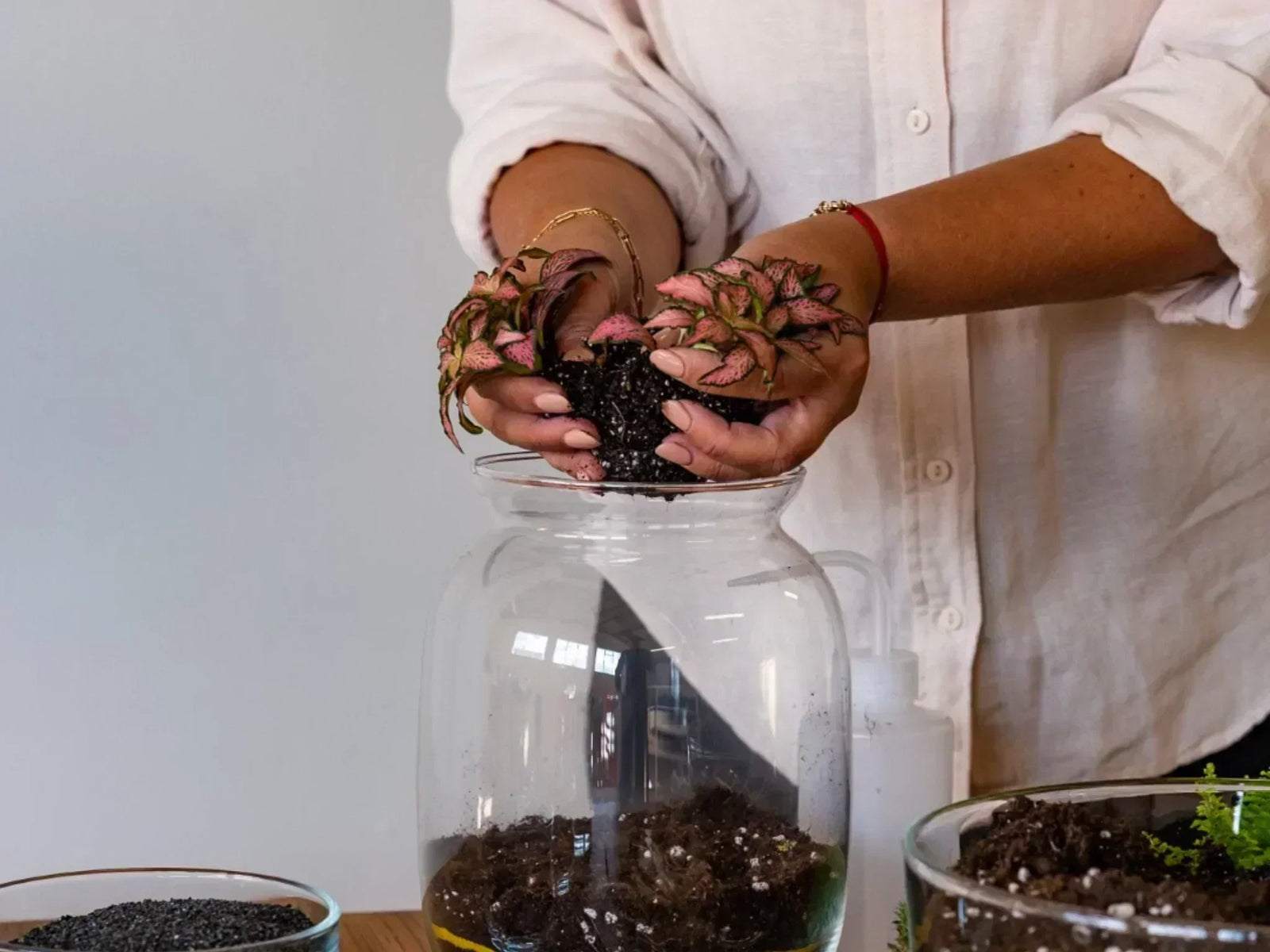 A person wearing a light-colored shirt is carefully placing a small plant with pink-tipped leaves into a glass terrarium. Surrounding them are various containers holding soil and decorative materials, creating a vibrant workshop setting.