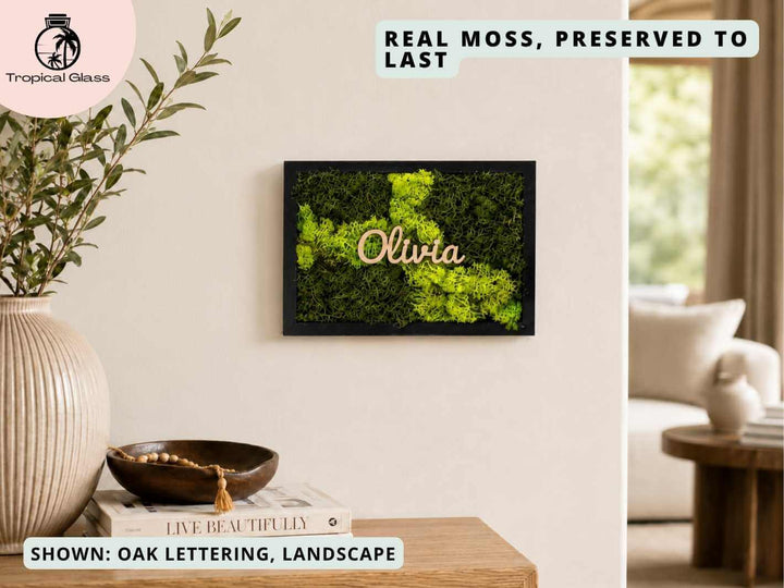 A personalized moss wall art frame featuring vibrant green preserved moss arranged in a decorative pattern, with the name Olivia in stylish oak lettering. The frame is displayed on a light-colored wall alongside a cozy living space.