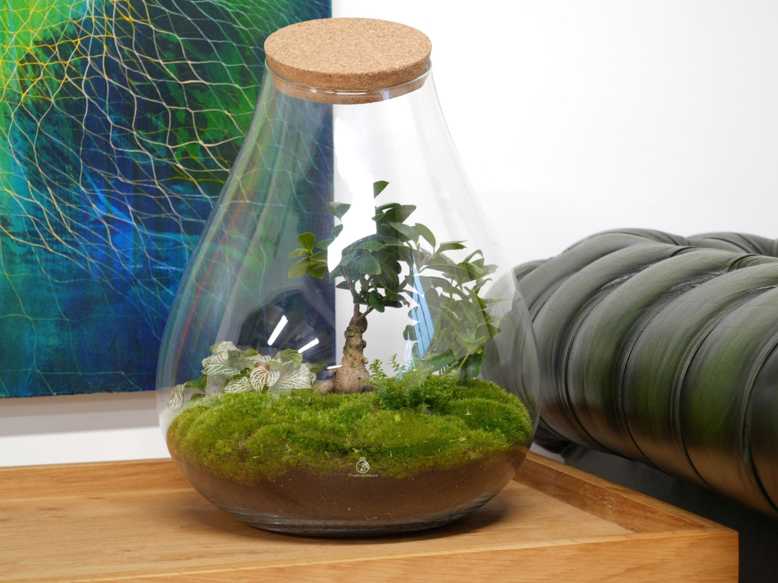 A teardrop-shaped glass terrarium sits on a wooden surface, showcasing a vibrant green landscape with various plants, including a Bonsai tree and lush moss.