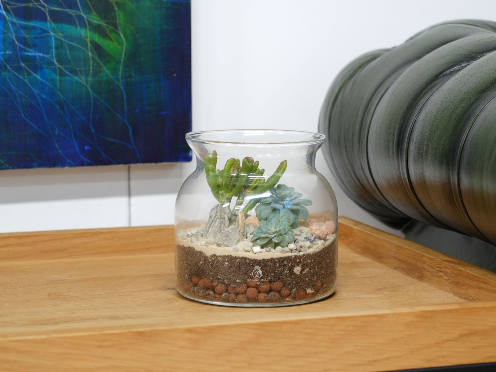 A glass terrarium sits on a wooden table, featuring a variety of succulent plants arranged with pebbles and soil. The backdrop includes a colorful abstract painting and a green upholstered chair, adding a modern touch to the serene scene.