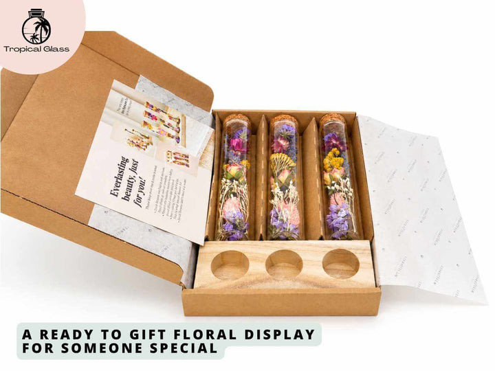 An open cardboard gift box reveals three glass tubes filled with an assortment of colorful dried flowers. The packaging includes a decorative insert and a wooden stand with circular cutouts for displaying the floral tubes elegantly.