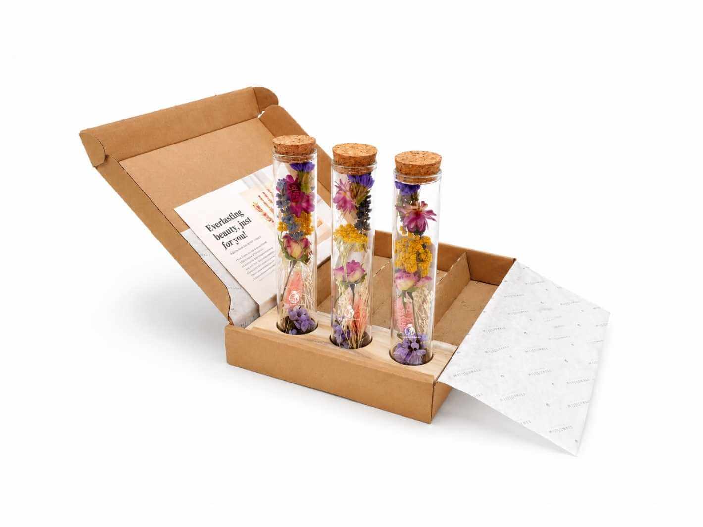 A gift set featuring three glass tubes filled with vibrant dried flowers, each uniquely arranged, sits in a brown cardboard box with a cork stopper on each tube. The box is partially open, revealing an accompanying card inside.