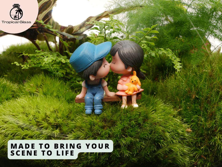 A charming mini couple figurine sits on a wooden bench, sharing a sweet kiss while surrounded by lush green moss and plants.