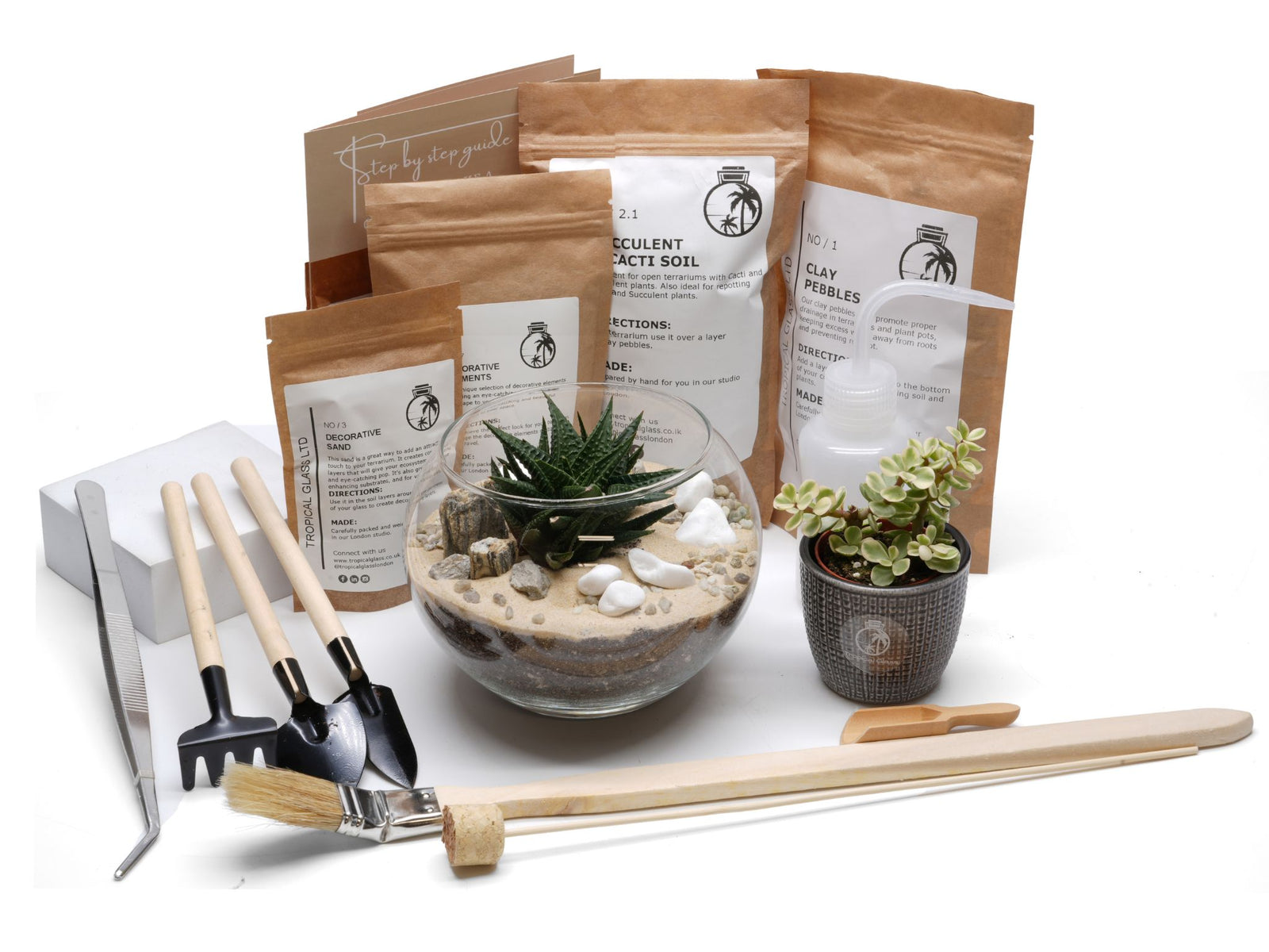 A complete DIY terrarium kit is displayed, featuring a glass container with a layered sandy base, green succulents, and decorative pebbles. The kit includes various soil and care supplies, along with tools for assembling the terrarium.