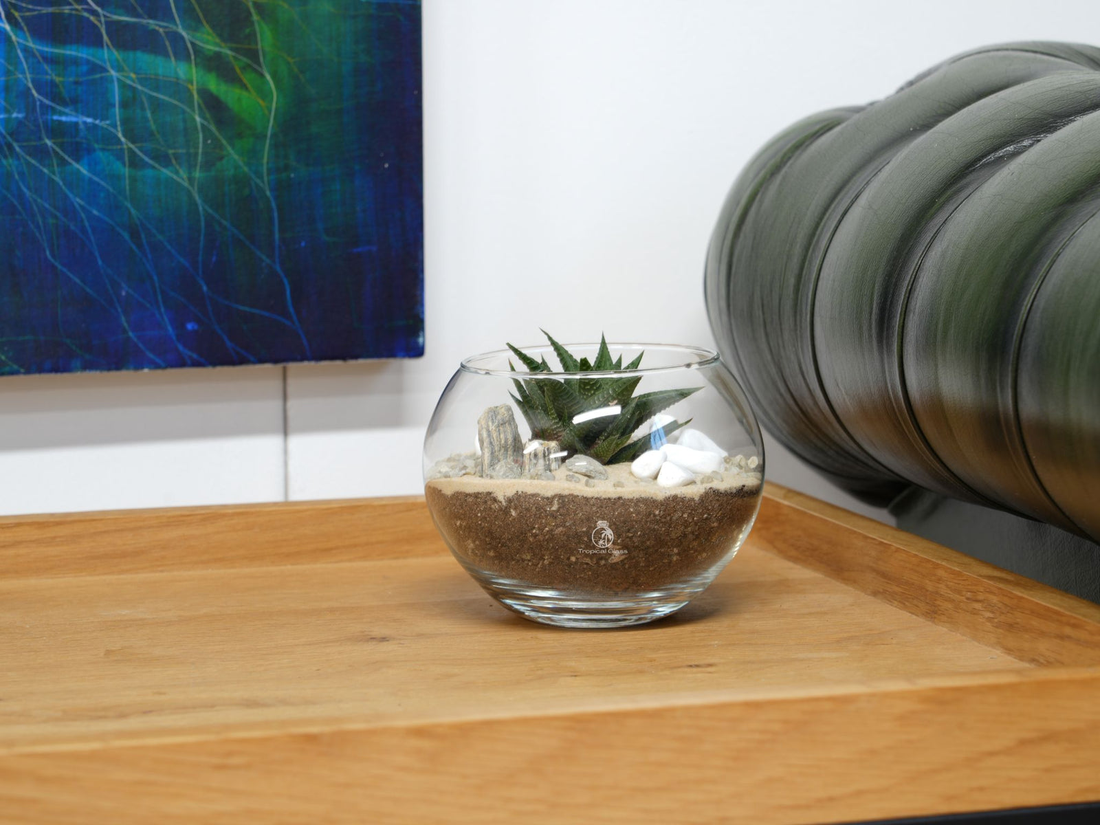 A round glass terrarium sits on a wooden tray, featuring a small succulent surrounded by sand and decorative stones. The background includes a colorful abstract painting and a tufted green leather couch, adding to the cozy atmosphere.