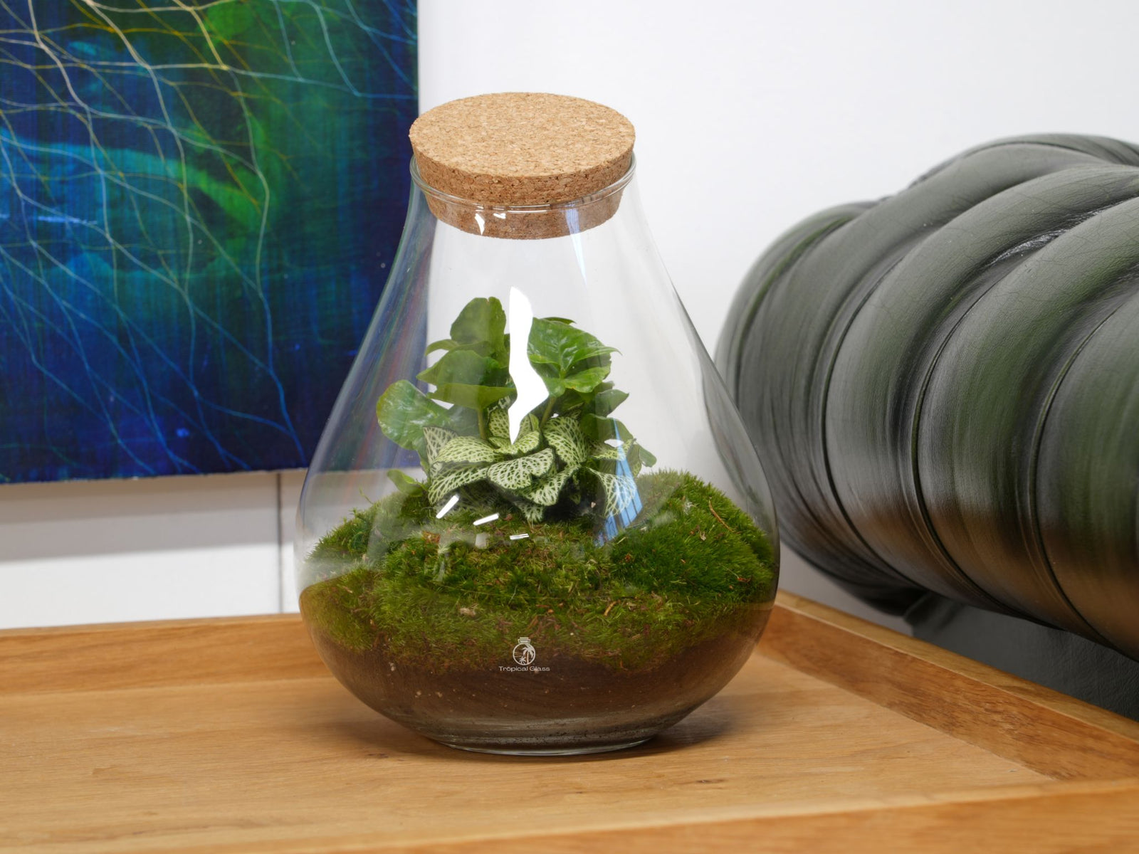 A stylish glass terrarium featuring lush green plants, including a small leafy plant and moss, sits on a wooden table. The terrarium is topped with a cork lid and surrounded by a colorful abstract painting and a green upholstered chair.