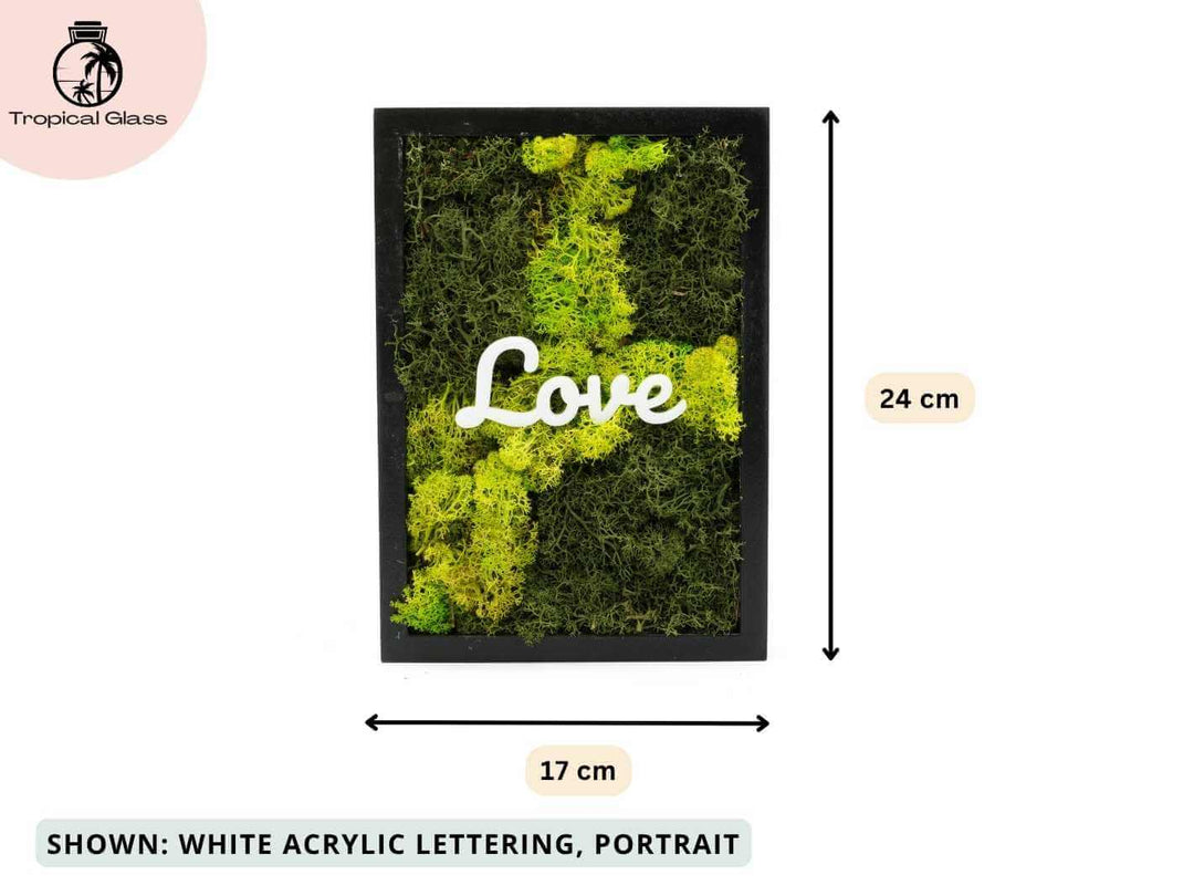 A rectangular black frame displays vibrant preserved moss in shades of green, with the word Love elegantly crafted in white acrylic lettering at the center.