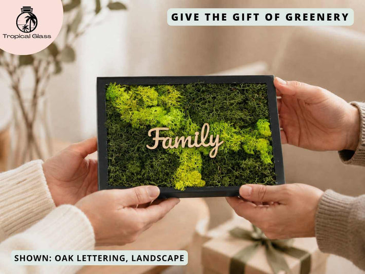 A person holds a personalized moss wall art frame featuring vibrant green moss and the word Family in elegant oak lettering. The frame is surrounded by a cozy, indoor setting with soft textiles and plants in the background.