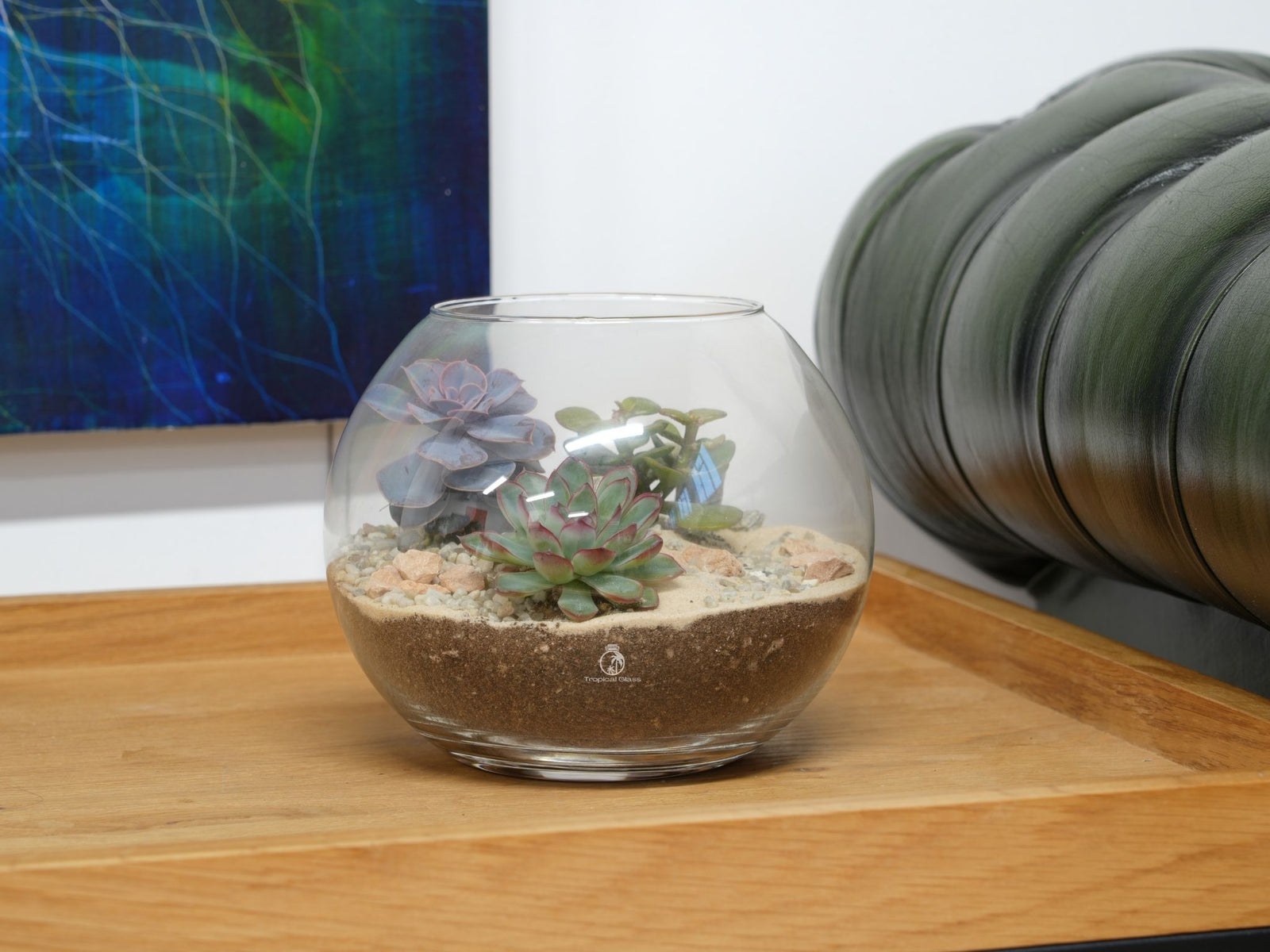 A clear glass fishbowl terrarium sits on a wooden surface, featuring an arrangement of colorful succulents and cacti nestled in sandy soil and decorative stones.