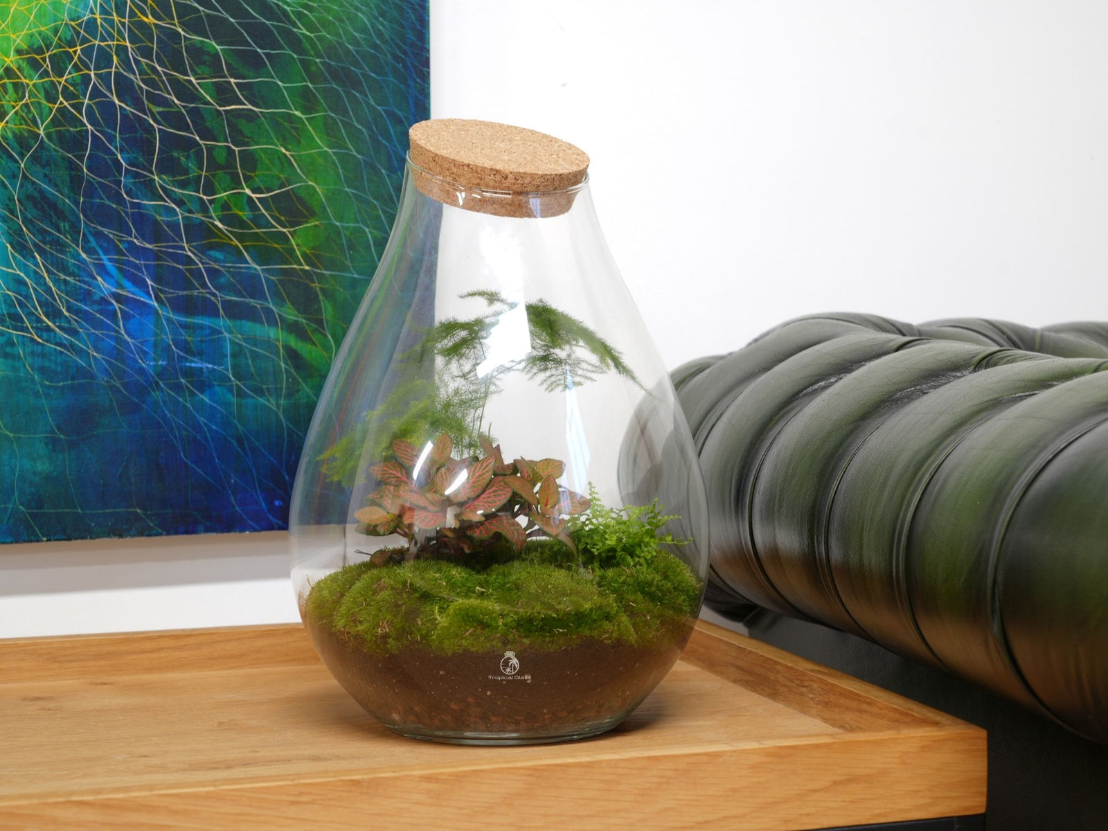 A teardrop-shaped glass terrarium sits on a wooden table, showcasing vibrant green moss and various plants, including Fittonia and ferns, beneath a cork lid. The background features an abstract artwork and a dark green leather sofa.