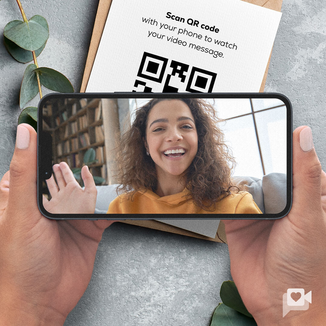 A person with curly hair smiles and waves while holding a smartphone in their hands, which displays their video message. The background shows a cozy living space, and a gift box with a QR code is placed nearby, inviting viewers to scan it.