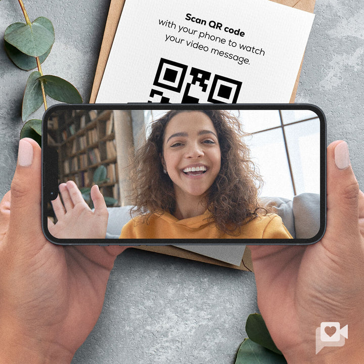 A person with curly hair smiles and waves while holding a smartphone in their hands, which displays their video message. The background shows a cozy living space, and a gift box with a QR code is placed nearby, inviting viewers to scan it.