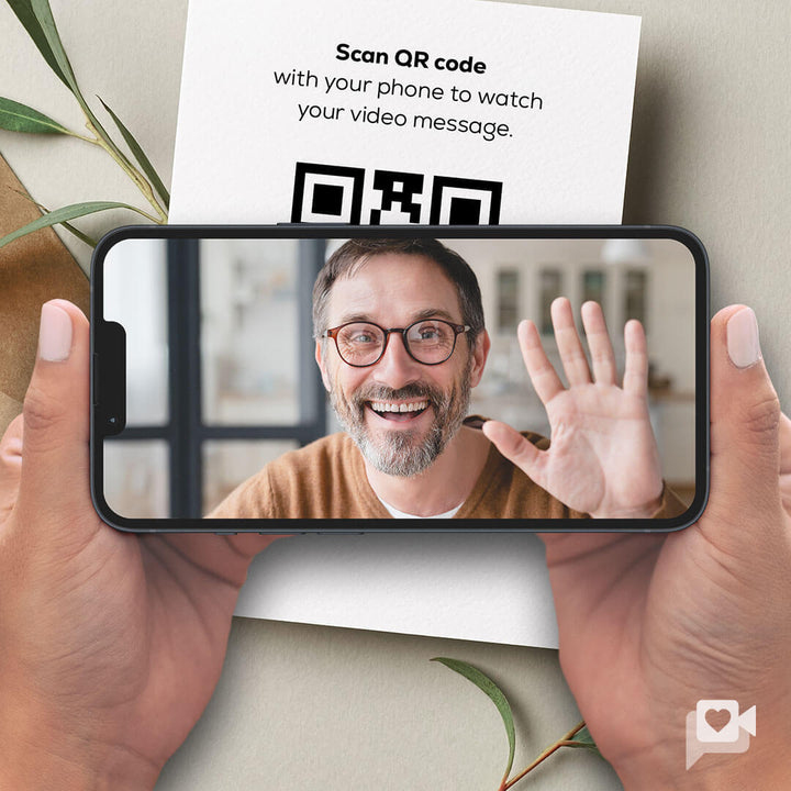 A person with glasses smiles and waves while holding a smartphone displaying their video message. In the background, a QR code and text inviting viewers to scan it are visible on a piece of paper, surrounded by green leaves.