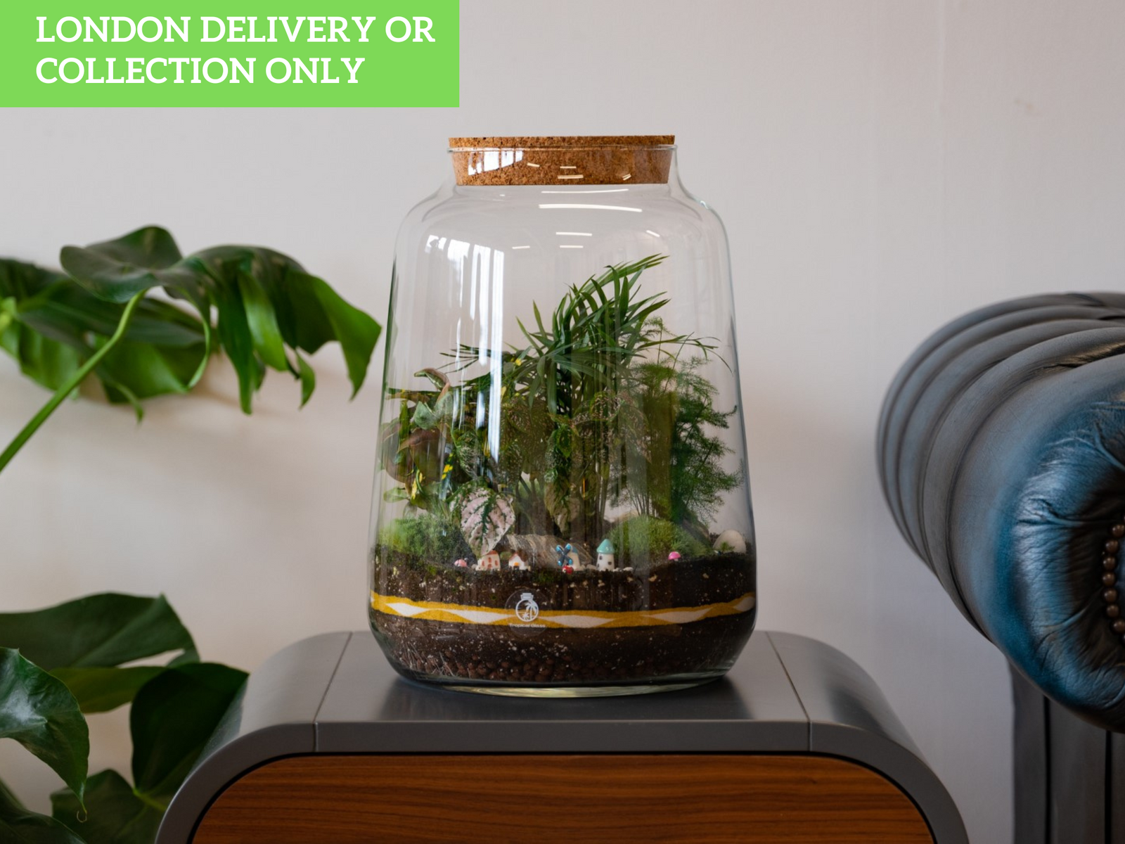 A glass terrarium with a cork lid sits atop a wooden table, showcasing a lush arrangement of greenery, including various plants and decorative elements. A green sign in the upper left corner indicates LONDON DELIVERY OR COLLECTION ONLY.