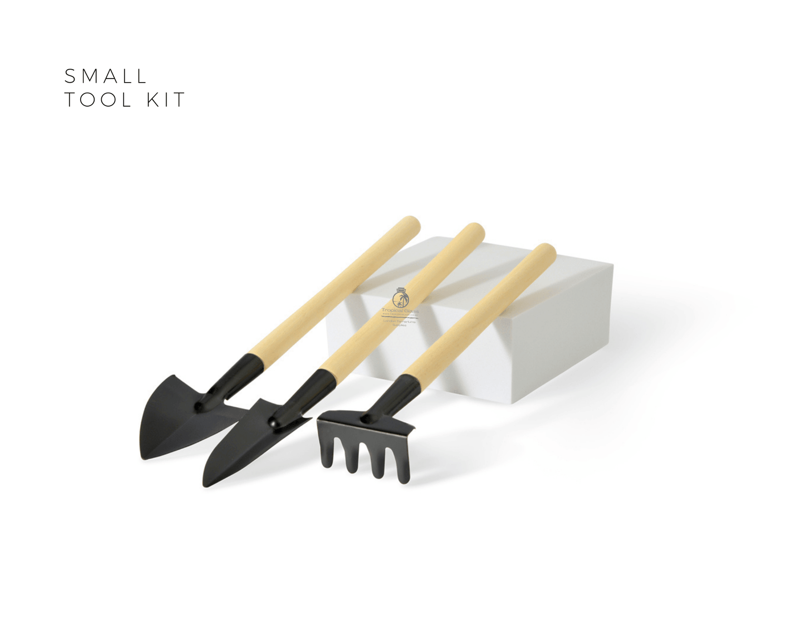 A small garden tool kit featuring three metal tools with wooden handles, including a spade, a shovel, and a rake, arranged neatly beside a white box. The tools are designed for indoor gardening and terrarium creation.