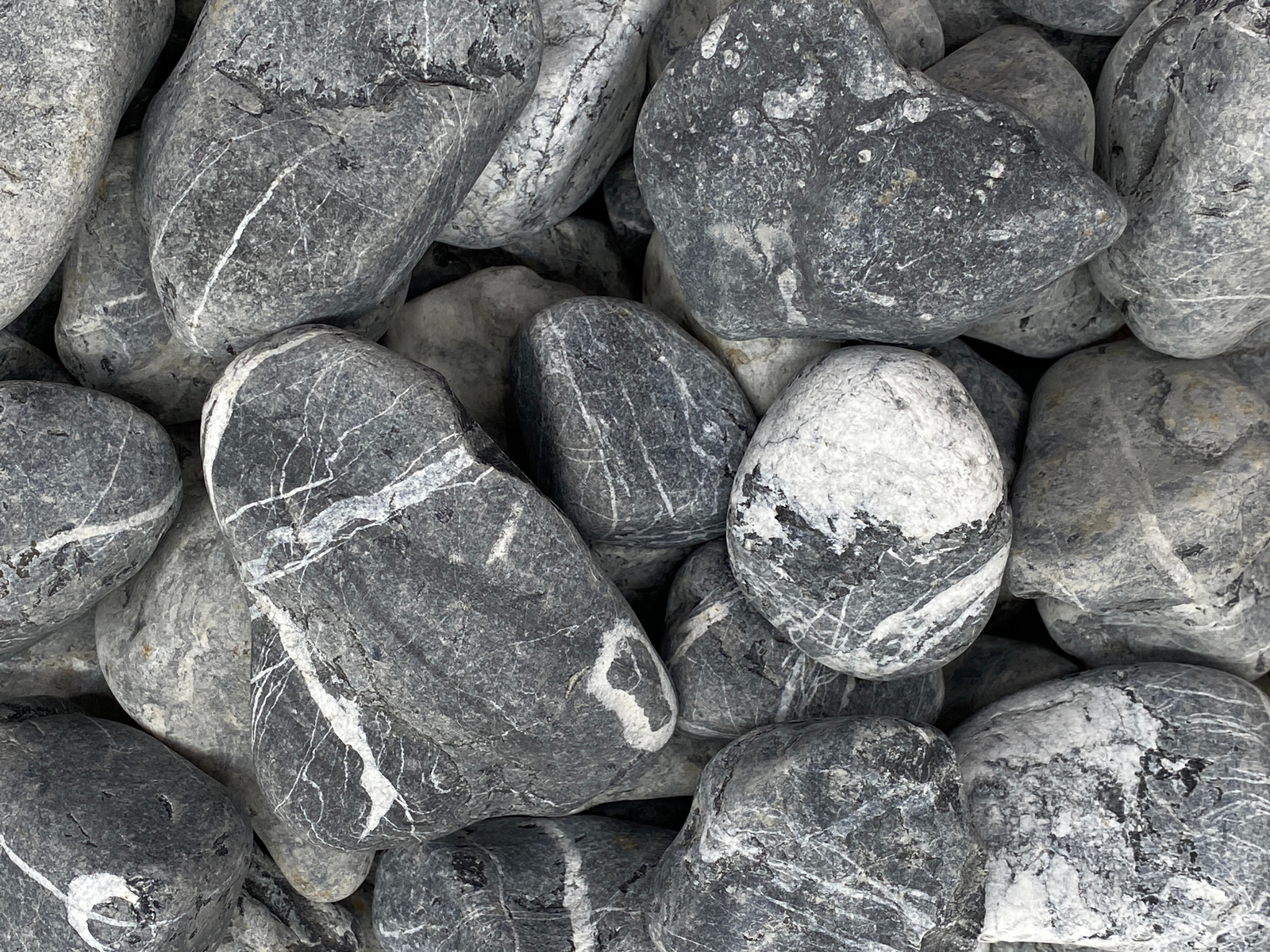 A close-up view of a collection of natural black and gray pebbles, varying in size from 4 to 10 cm. The pebbles feature smooth surfaces and intricate veining, creating a visually striking texture suitable for decorative use.