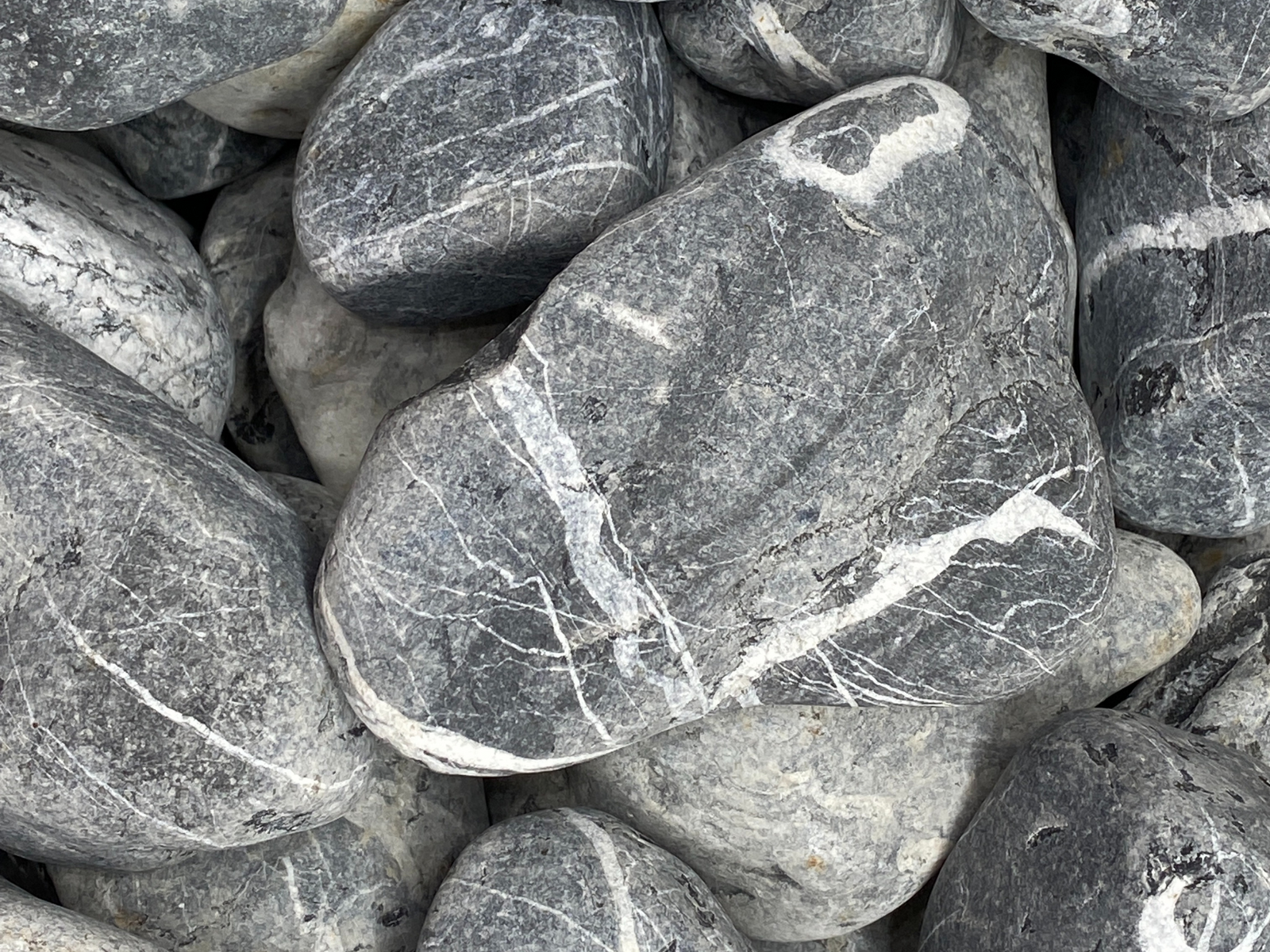 A close-up view of natural black and gray pebbles, showcasing their smooth surfaces and varied textures. The pebbles range in size and feature striking white veins running through the darker stone, creating a visually appealing contrast.