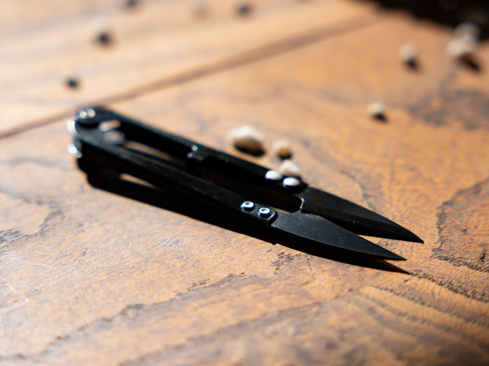 A pair of black pruning snips is positioned on a wooden surface, showcasing its sharp, pointed blades and compact design. Surrounding the snips are small plant seeds, indicating a gardening or plant care context.