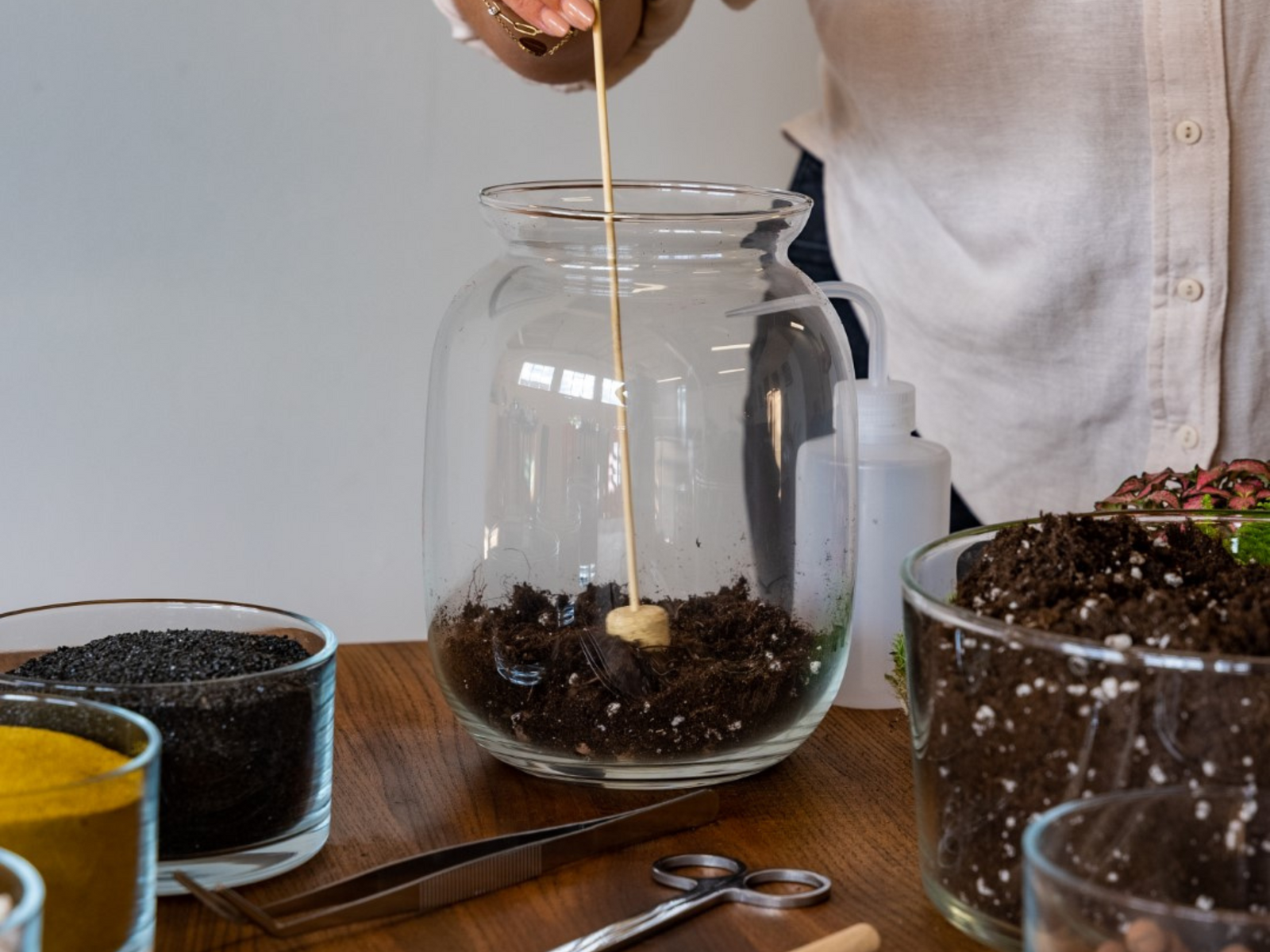 A person gently presses a cork tamper into soil inside a glass terrarium, surrounded by various bowls of colorful materials like black gravel and yellow sand.