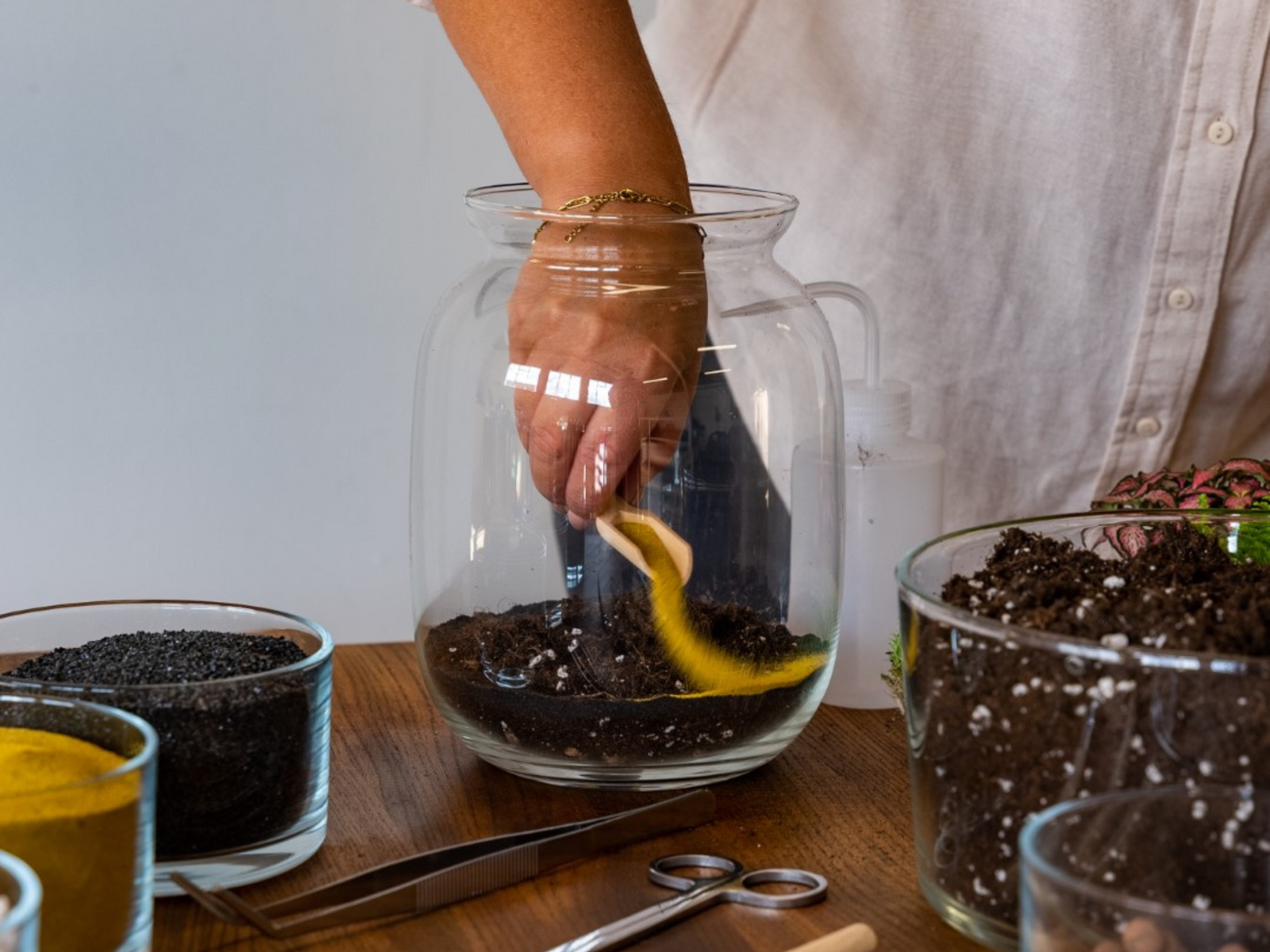 A person is using a small wooden scoop to add yellow sand into a glass jar filled with soil. Surrounding the jar are various bowls containing different materials, including black gravel and colorful sand, on a wooden table.
