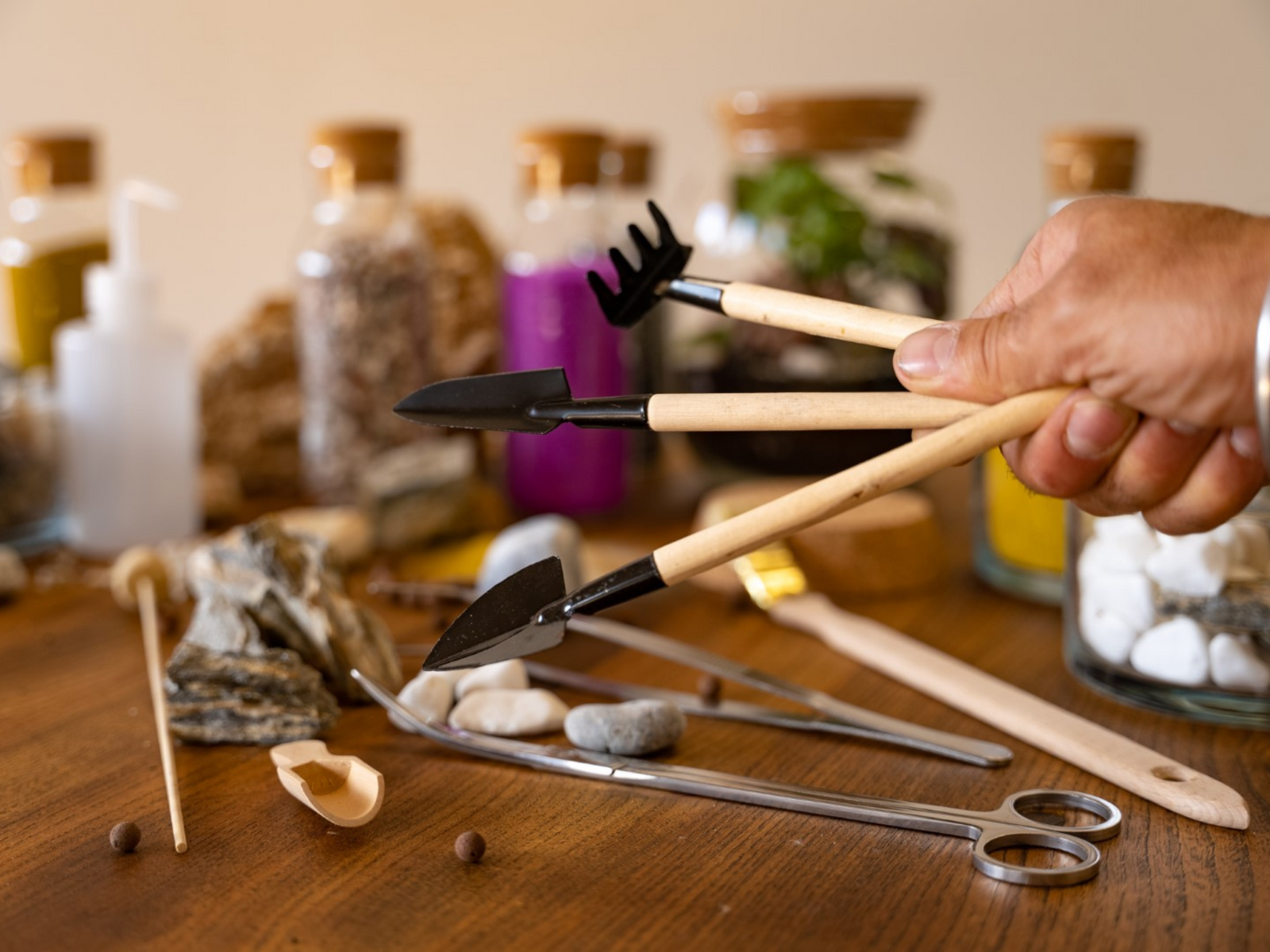 A hand holds three small gardening tools, including a spade, rake, and shovel, above a wooden table scattered with pebbles, decorative rocks, and various jars filled with colorful materials.